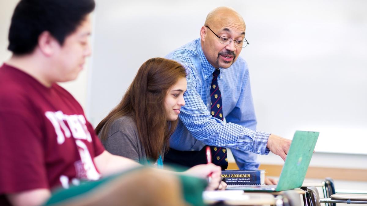 professor billy burke stands near 2 students in a classroom, pointing at the laptop screen for one of them